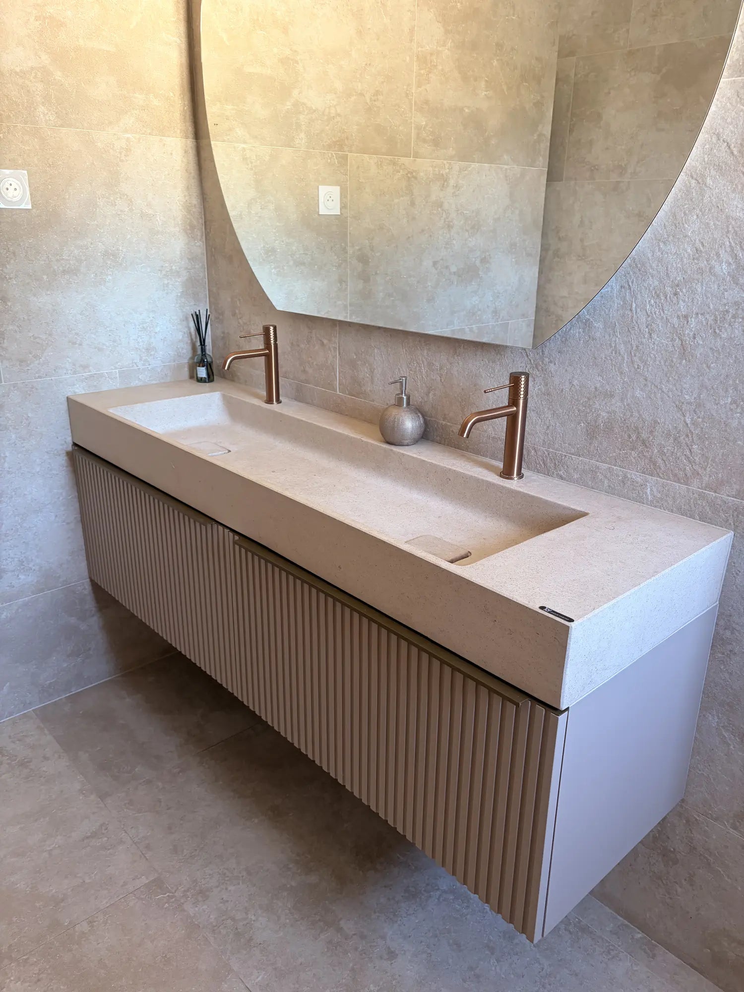Plan de vasque double minimaliste en pierre naturelle crème Moléanos, deux cuves intégrées avec bonde Magique carrée en pierre, robinetterie cuivrée, meuble suspendu cannelé beige, grand miroir rond, salle de bain contemporaine en travertin
