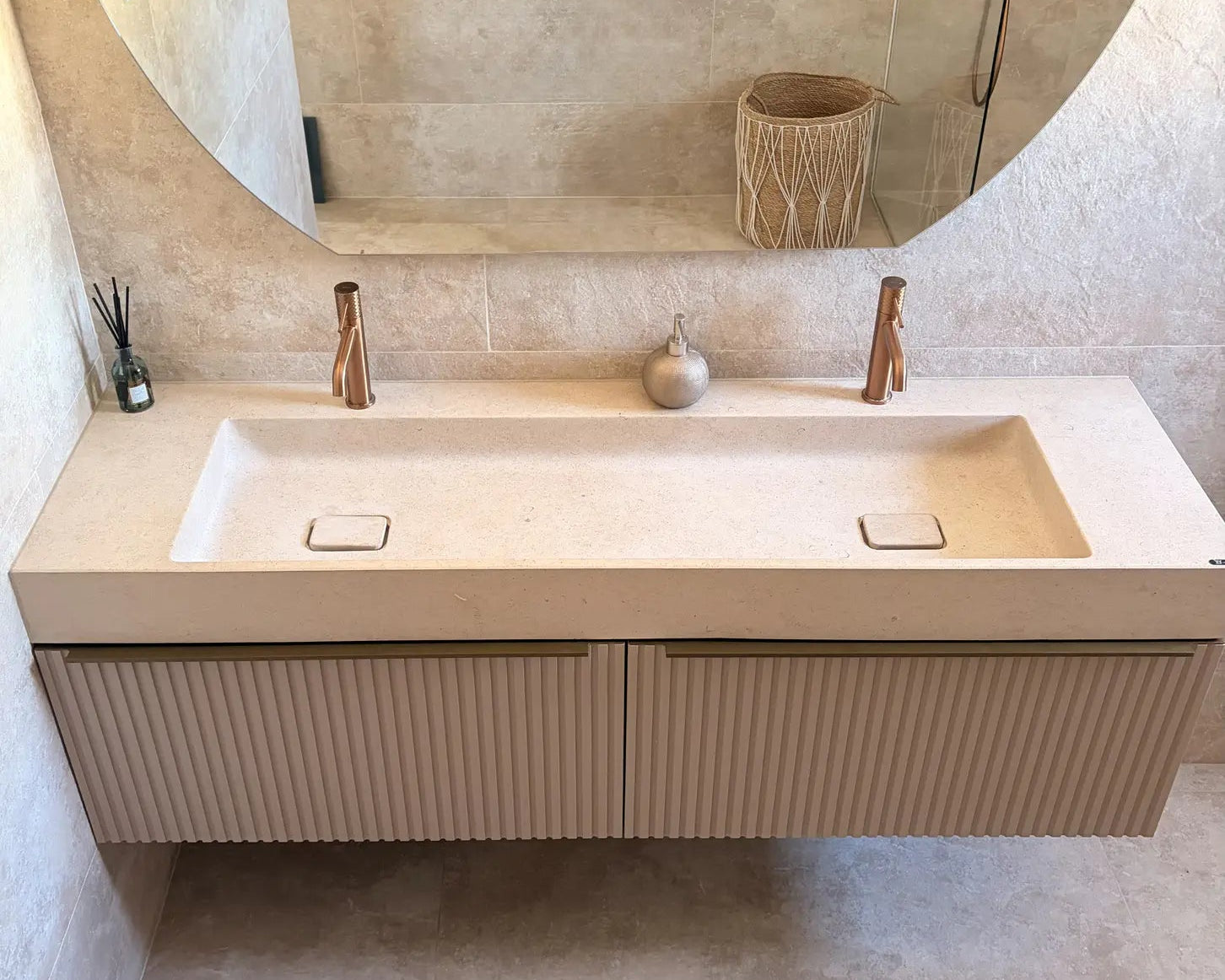 Plan de vasque double minimaliste en pierre naturelle crème Moléanos, deux cuves intégrées avec bonde Magique carrée en pierre, robinetterie cuivrée, meuble suspendu cannelé beige, grand miroir rond, salle de bain contemporaine en tons crème