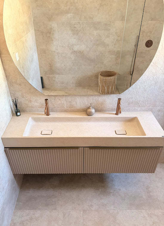 Plan vasque en pierre beige naturelle crème Moléanos, double cuve intégrée suspendue, bonde Magique en pierre, meuble cannelé, robinetterie cuivre, grand miroir rond, salle de bain contemporaine
