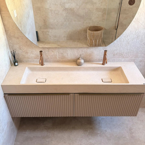 Plan vasque en pierre beige naturelle crème Moléanos, double cuve intégrée suspendue, bonde Magique en pierre, meuble cannelé, robinetterie cuivre, grand miroir rond, salle de bain contemporaine