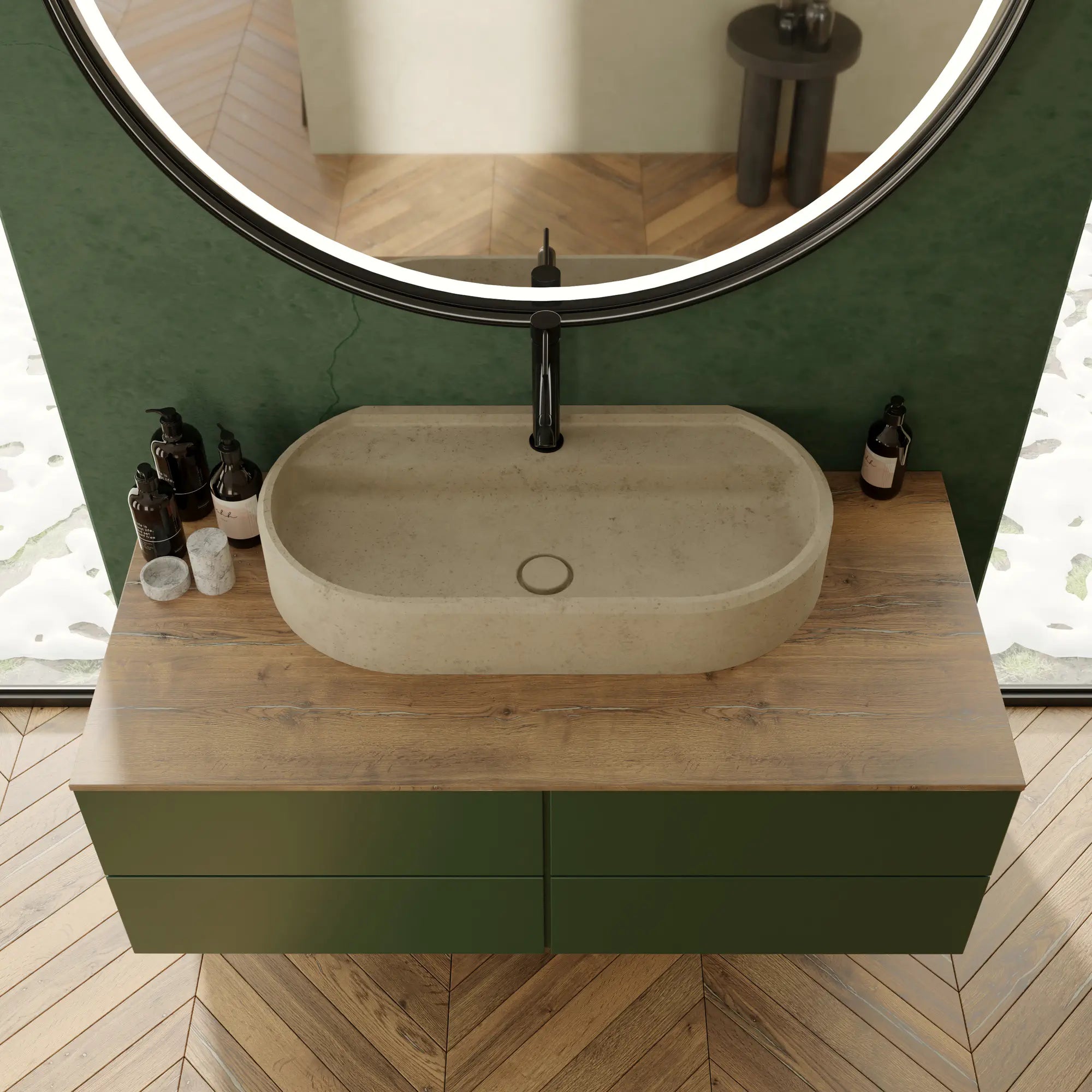 Vasque à poser ovale en pierre naturelle beige crème Moléanos, posée sur un meuble en bois et vert, robinetterie noire, grand miroir rond lumineux, mur vert, parquet chevron, salle de bain contemporaine