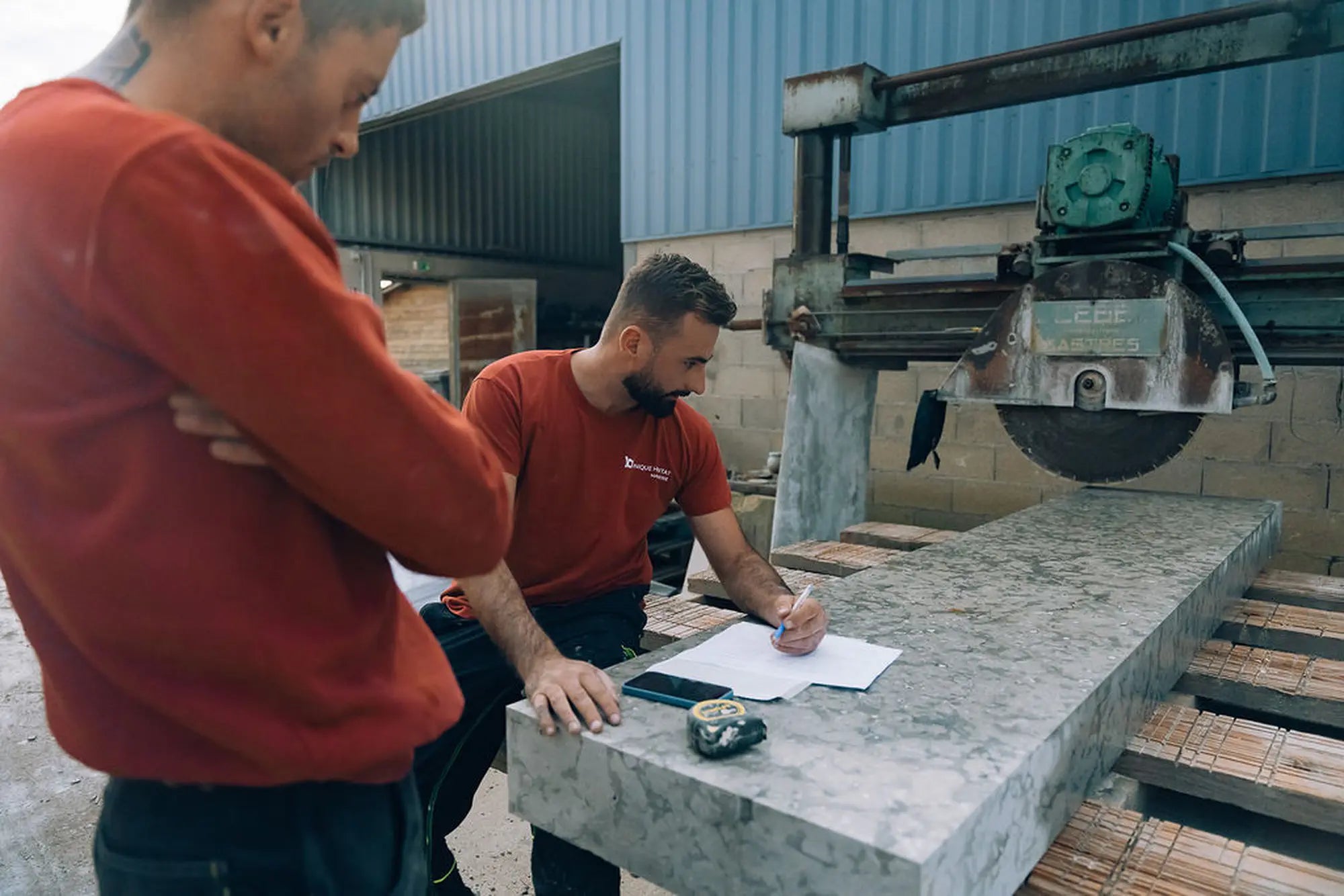 Artisans Unique Habitat planifiant un projet de vasque suspendue en pierre sur-mesure dans l'atelier, prise de mesures et plan sur un bloc de pierre, scie à pierre, fabrication française


