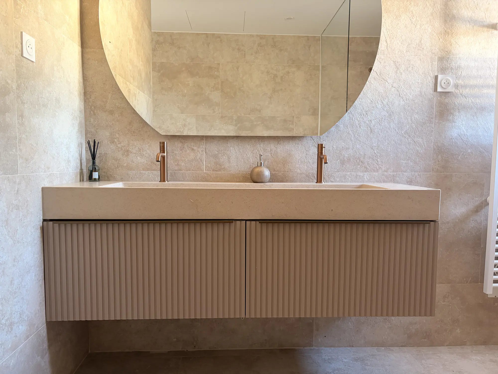 Vasque suspendue en pierre naturelle crème Moléanos, double cuve, meuble cannelé, robinetterie cuivre, grand miroir rond, salle de bain contemporaine