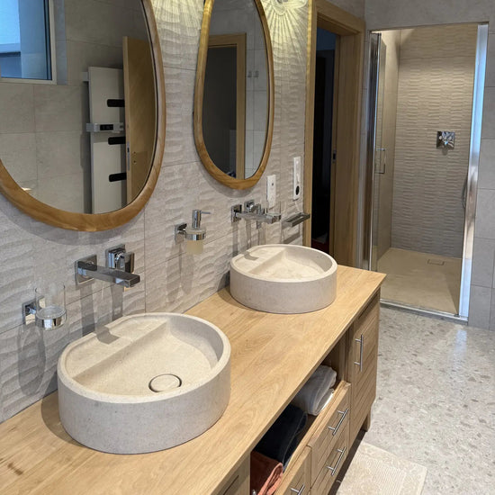 Salle de bain avec double vasque à poser ronde en pierre crème sur un plan en bois, miroirs ovales à cadre en bois et robinetterie murale