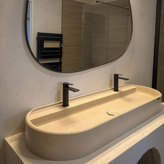Vasque ovale à poser Salagou de 120 cm en pierre crème de Moléanos, avec deux robinets noirs et un miroir de forme organique, sur un meuble en béton ciré à arche